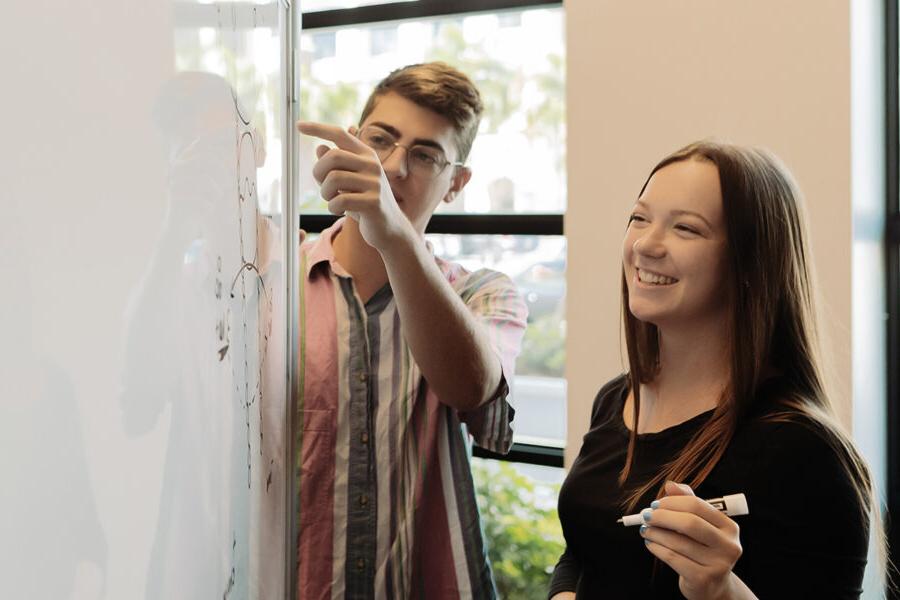 Two PBA students draw diagrams on a whiteboard.